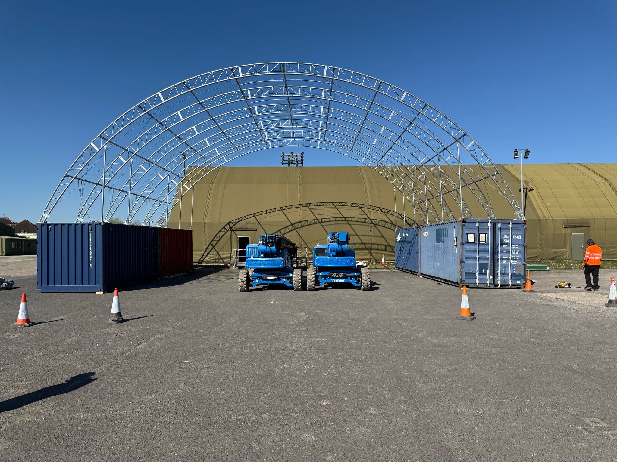 Steel framework of a SkyShield container canopy being assembled between two shipping containers with construction workers on site.